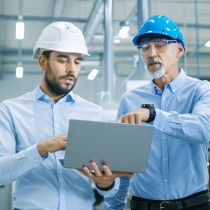 Zwei Männer mit Arbeitshelmen und Laptop diskutieren, während sie durch moderne Fabrik laufen