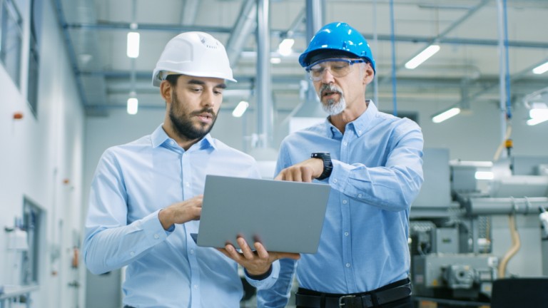 Zwei Männer mit Arbeitshelmen und Laptop diskutieren, während sie durch moderne Fabrik laufen